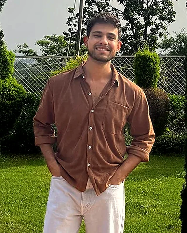 Aman smiling in a garden, hands in his pockets, wearing a brown shirt